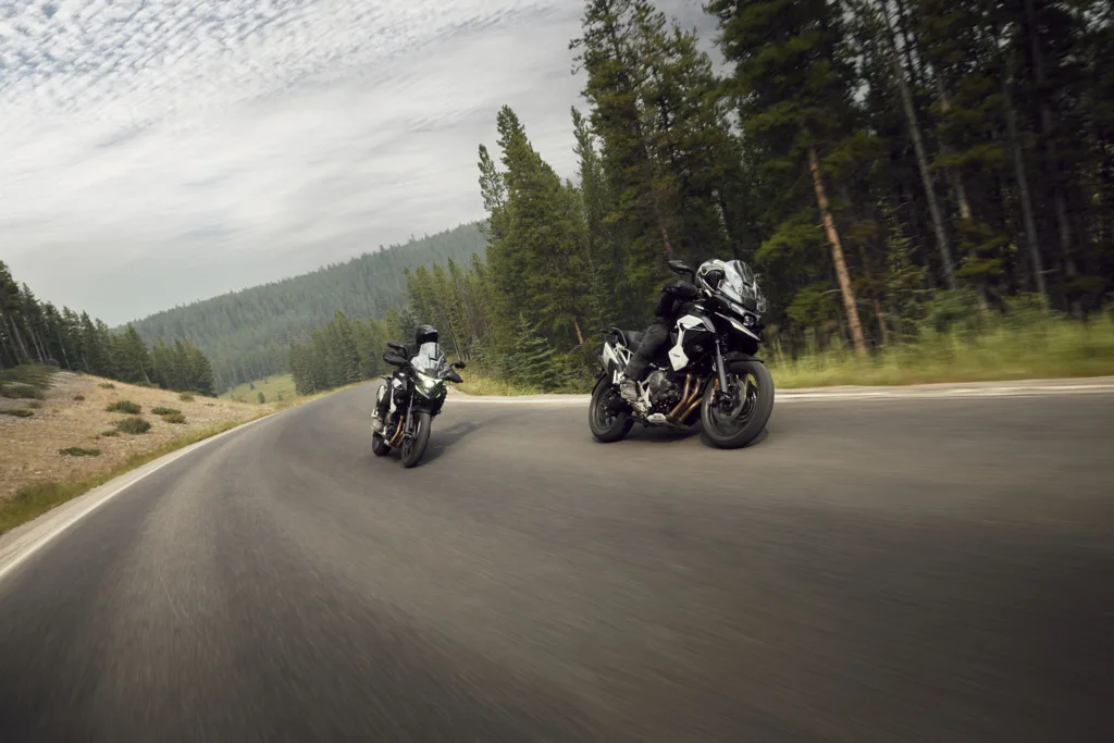 Two 2027 Triumph Tiger 1200 motorcycles being ridden on a forested mountain road.