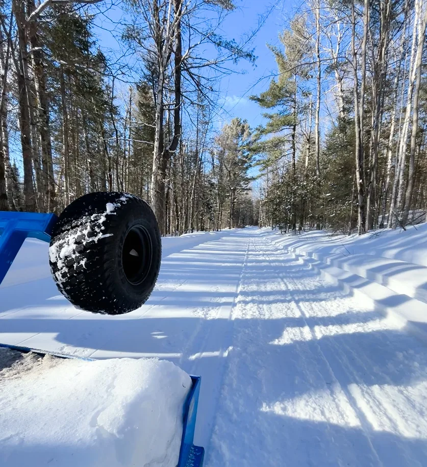 A groomed snowmobile trail through a sunlit forest, viewed from a vehicle.