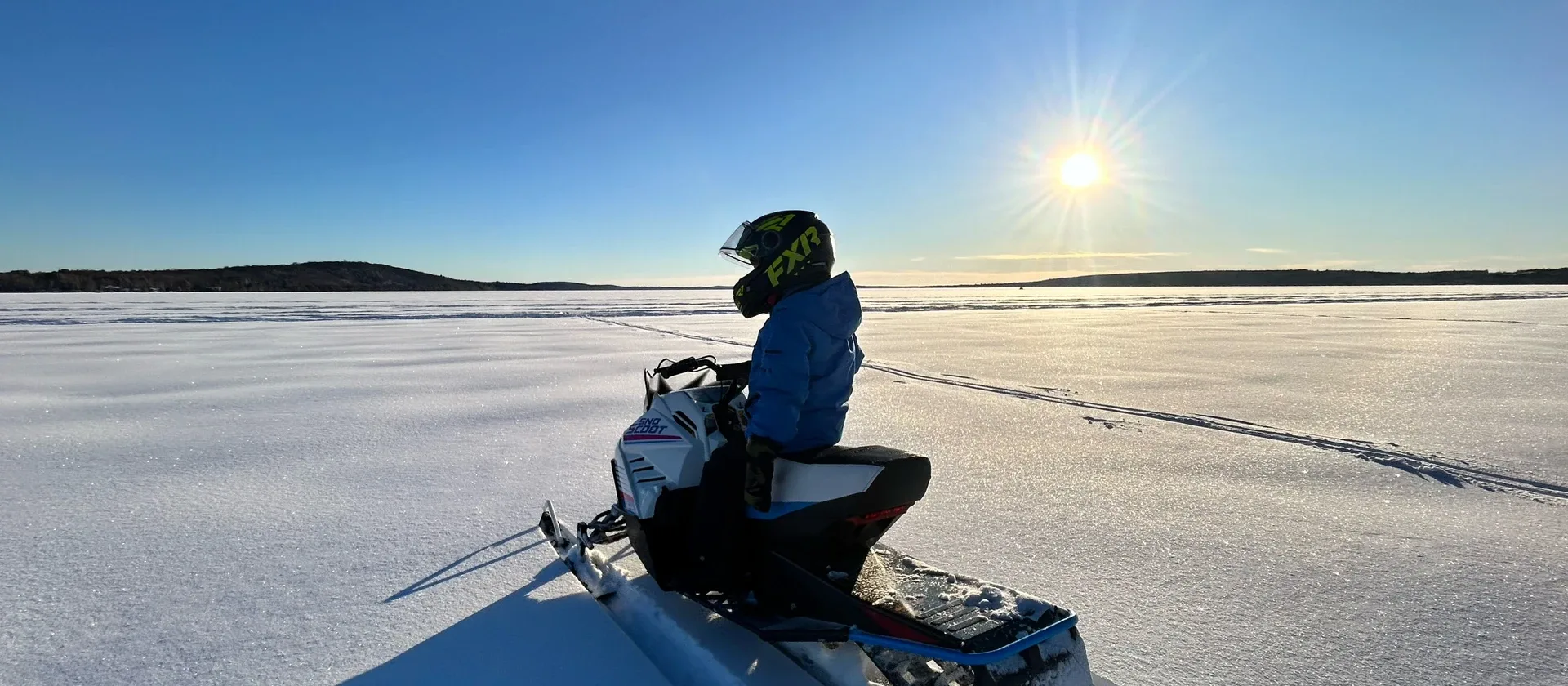 A snowmobiler seated on a sled in a wide snow-covered landscape beneath blue sky.